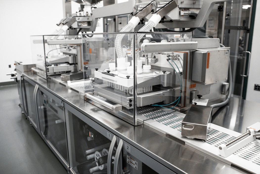An advanced pharmaceutical blister packaging machine, the Capital Investment Uhlmann B1440 Blister Line, captured in a sterile production environment. The machine, featuring robotic arms and a series of conveyors, is designed for precision in packaging tablets and capsules into blister packs. The image shows the intricate setup of the equipment with clear protective casing and operational controls, ready to handle the pharmaceutical packaging process.