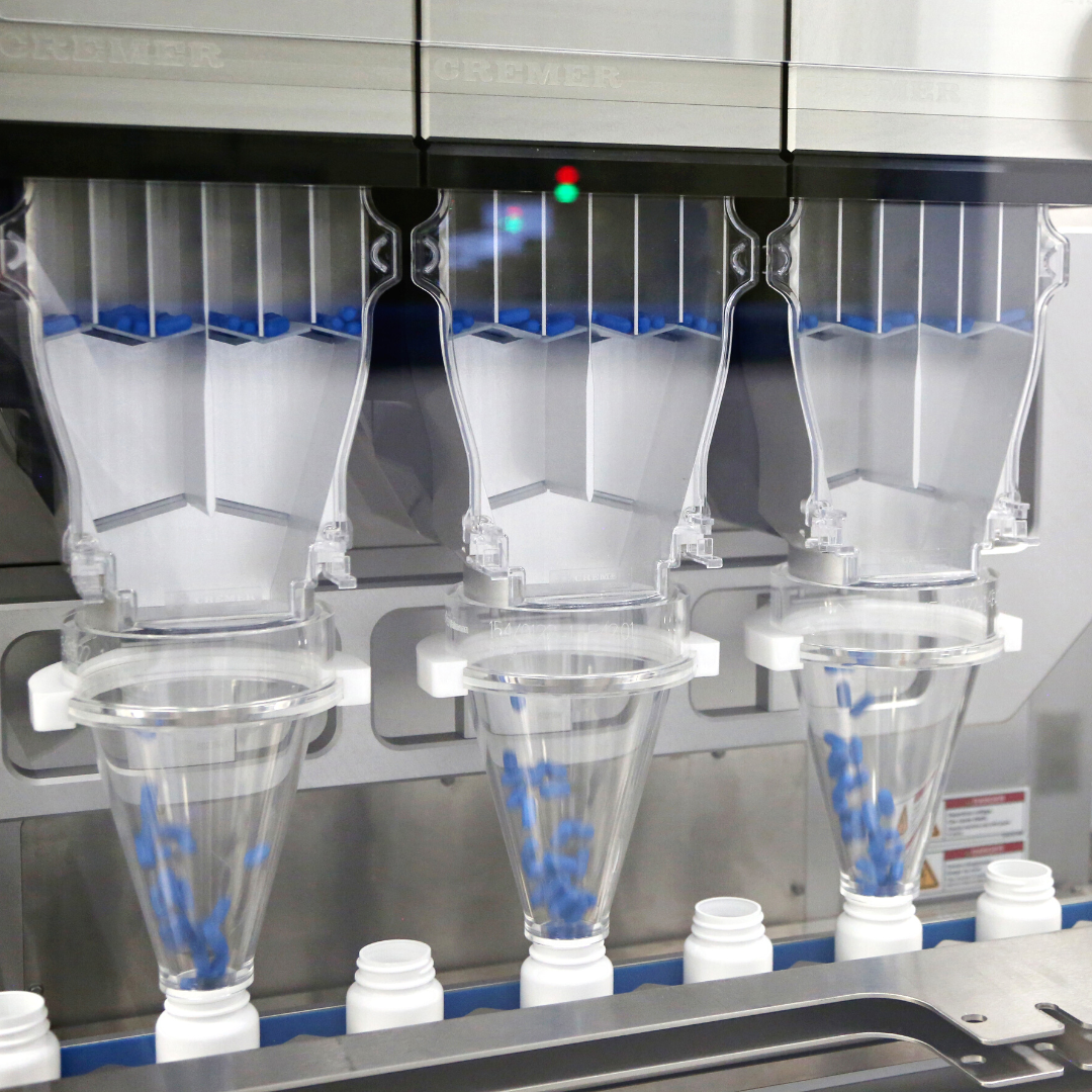 An automated pharmaceutical bottle packaging machine in operation, with clear channels filled with blue capsules, aligning and dispensing them into open bottles on a conveyor belt below. Visible in the frame is part of the machinery with the label "CREMER," and a control panel with indicator lights, suggesting precision and cleanliness in the pharmaceutical packaging process.