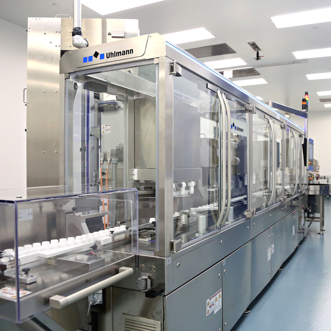 Image of a large, industrial Uhlmann IBC 150 pharmaceutical bottle packaging machine with clear protective panels. The machine, set in a clean, well-lit pharmaceutical manufacturing room, appears to be equipped with advanced technology for the automated packaging of medications into bottles.