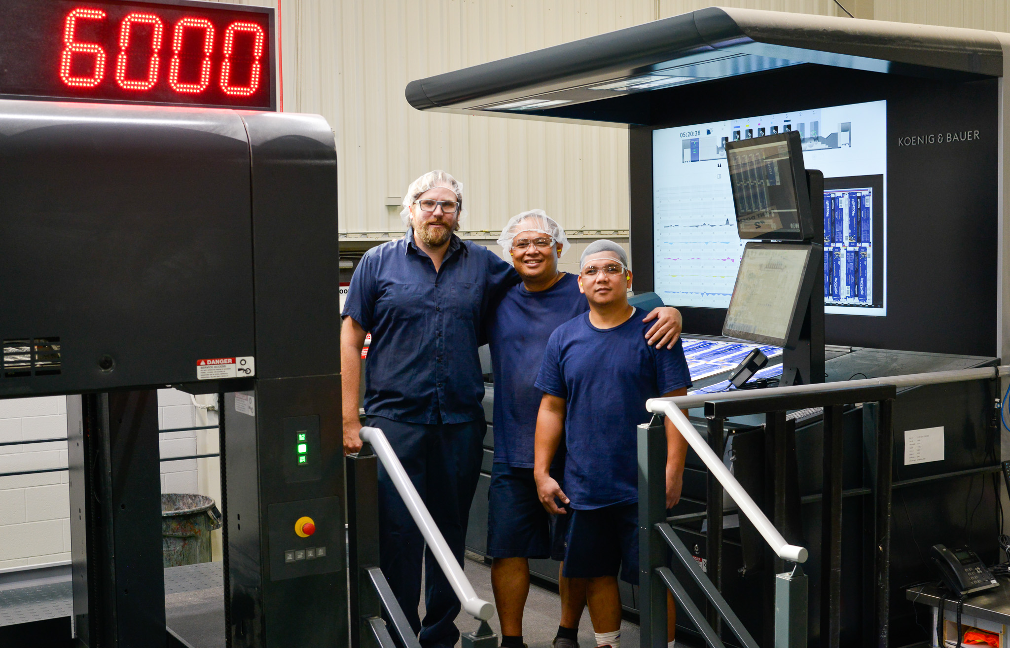 Three operators wearing blue shirts, safety glasses, and hairnets stand together, smiling beside the advanced Koenig & Bauer 7-Colour Litho Press in a production facility.